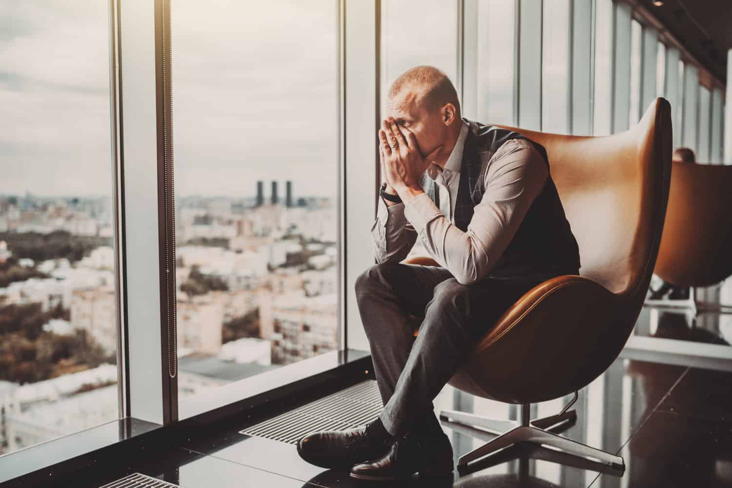 A stressed out adult caucasian man entrepreneur partly closing face with his hands while sitting on an office armchair next to the window of a modern business skyscraper and looking on the cityscape