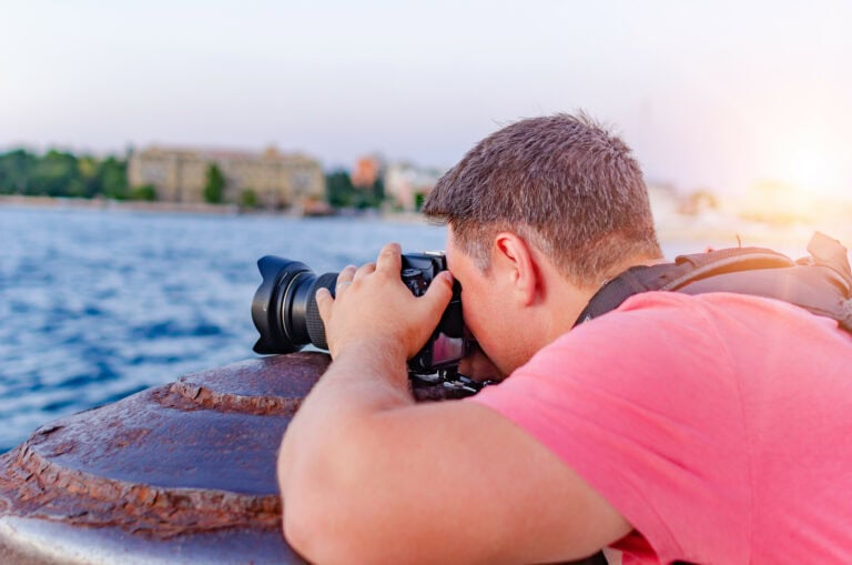 An outdoor enthusiast stands by the edge of a tranquil lake, framing a shot with his camera to capture the serene beauty of the water and surrounding landscape.
